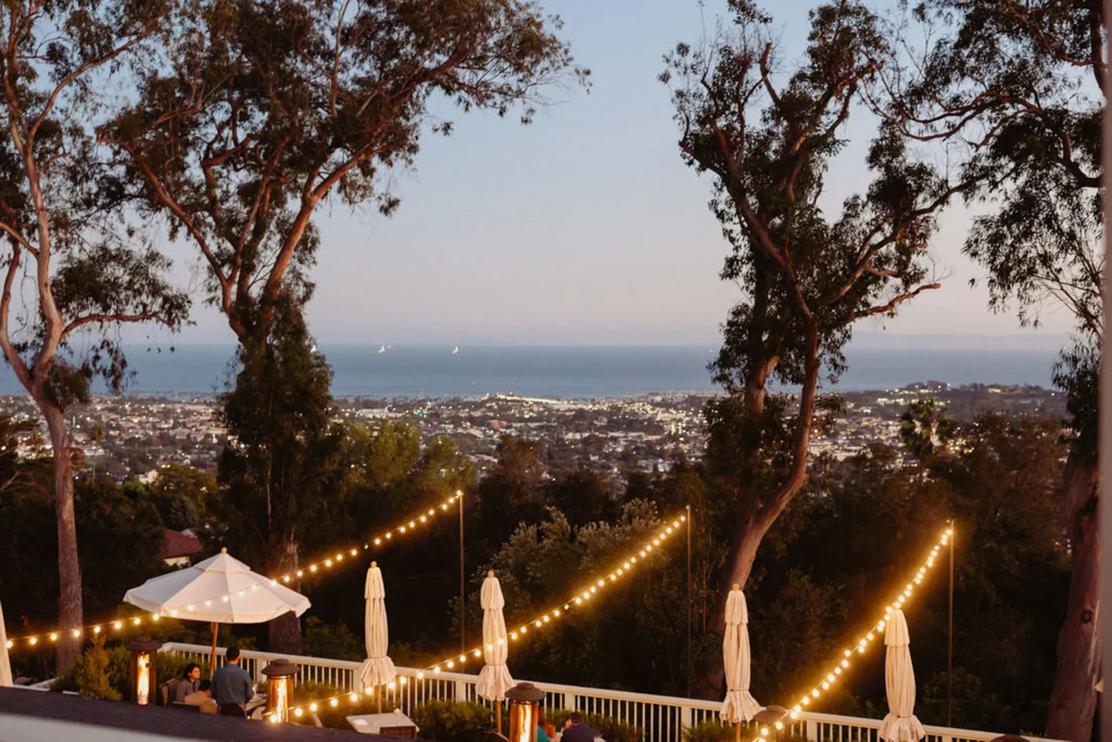 El Encanto's dining terrace at dusk, string lights strung between
                the oaks above the tables, with Santa Barbara and the ocean below