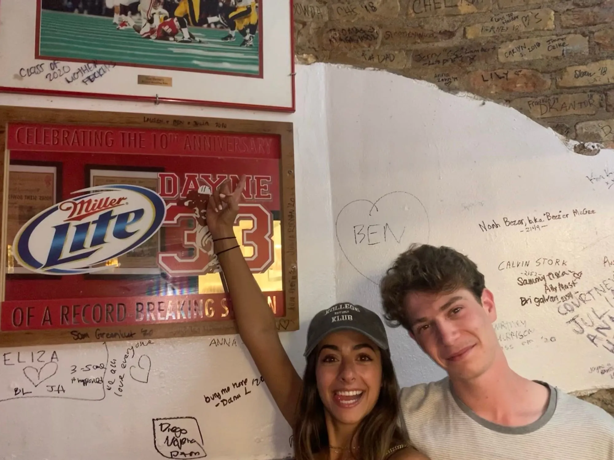 Lauren reaching up to a bar wall covered in handwritten signatures, Ben beside her