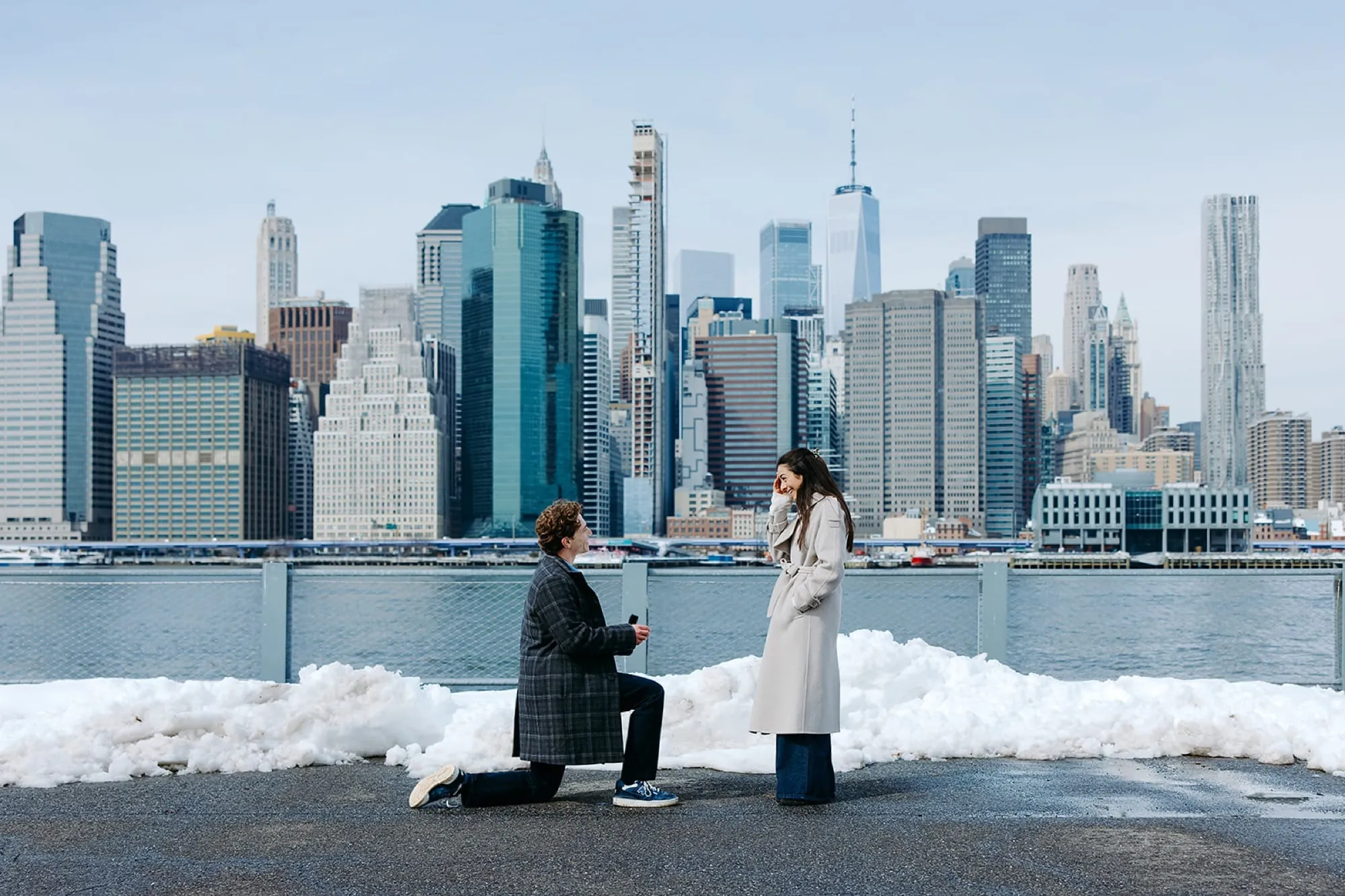Ben on one knee proposing to Lauren on the Brooklyn waterfront, the Manhattan skyline across the river