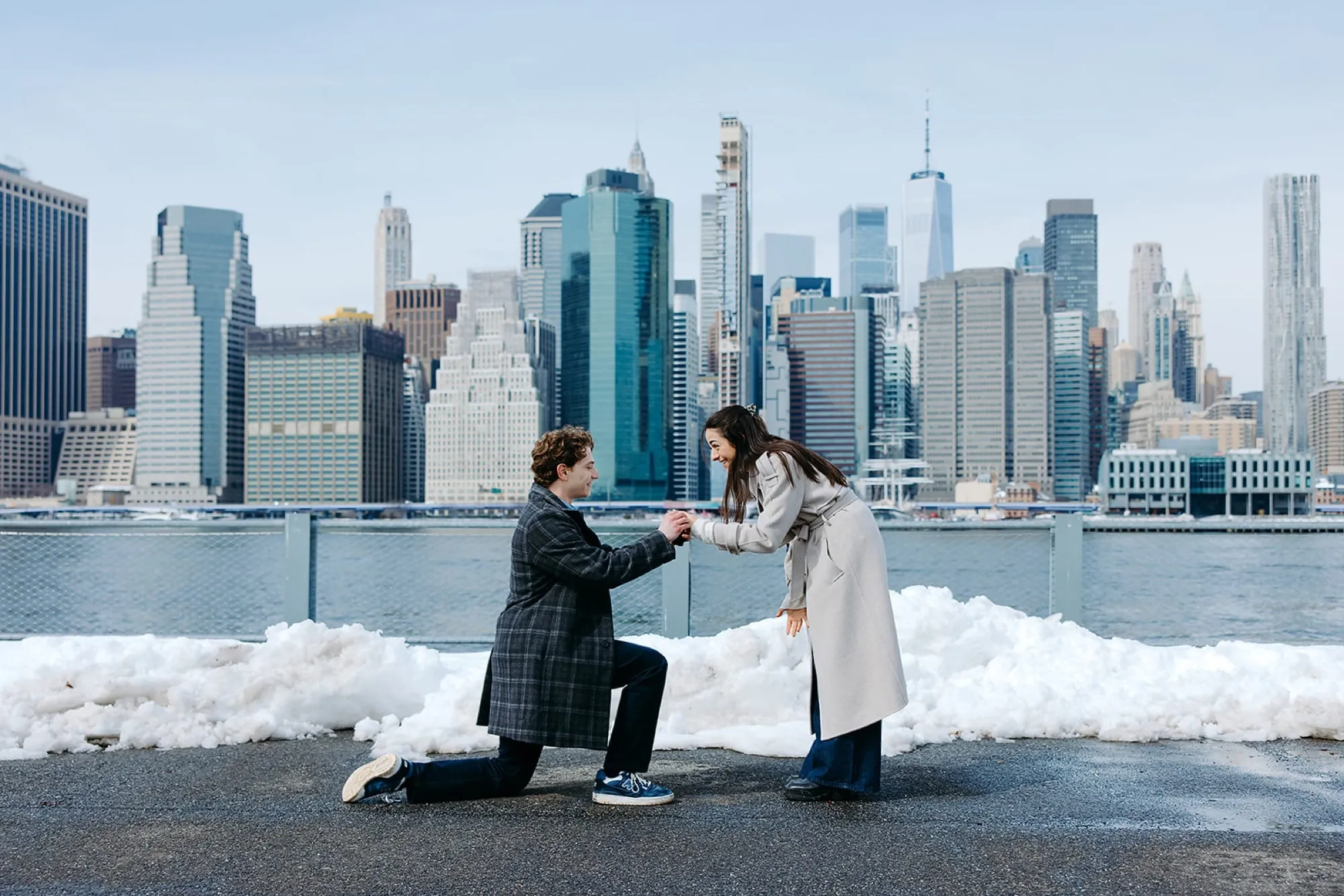 Ben on one knee taking Lauren’s hand, the Manhattan skyline across the water